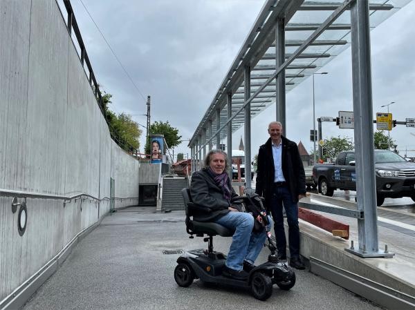 In einem elektrischen Rollstuhl sitzt Stephan Grumbt, Behindertenbeauftragter der Stadt Konstanz, und recht von ihm steht Bürgermeister Karl Langensteiner-Schönborn. Beide befinden sich vor Schräge zur Bushaltestelle am Sternenplatz.